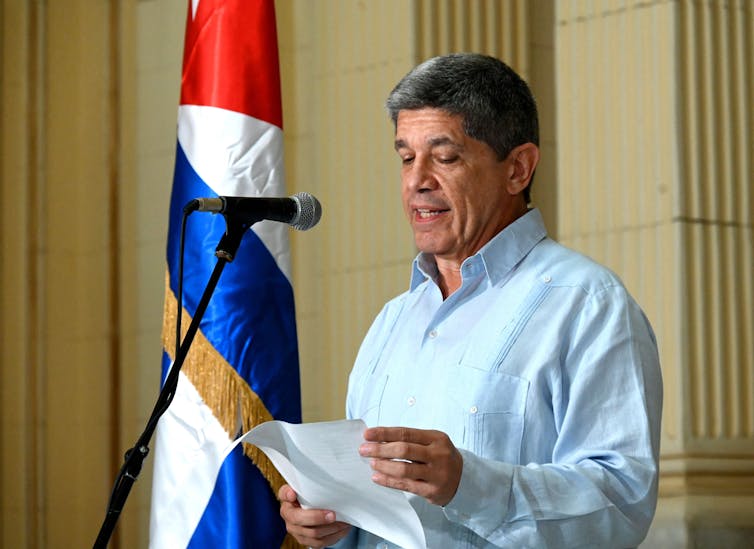A man wearing a light blue, open-collared shirt, reads into a microphone from a sheet of paper.