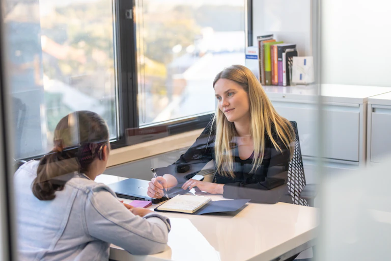 two women talk across desk. one wears a hearing device