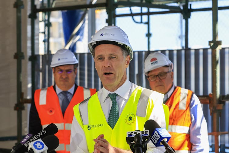 Man in high-vis vest and hard hat speaks into media microphones