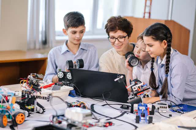 Una profesora rodeada de robots y aparatos tecnológicos les explica algo a sus alumnos en un ordenador portátil.