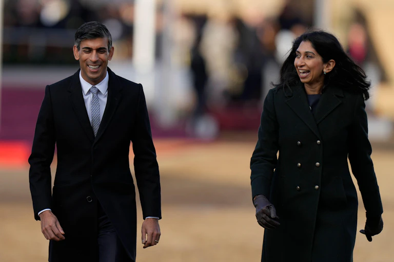 Rishi Sunak and Suella Braverman walking side by side, both smiling or laughing, in formal, black wool coats