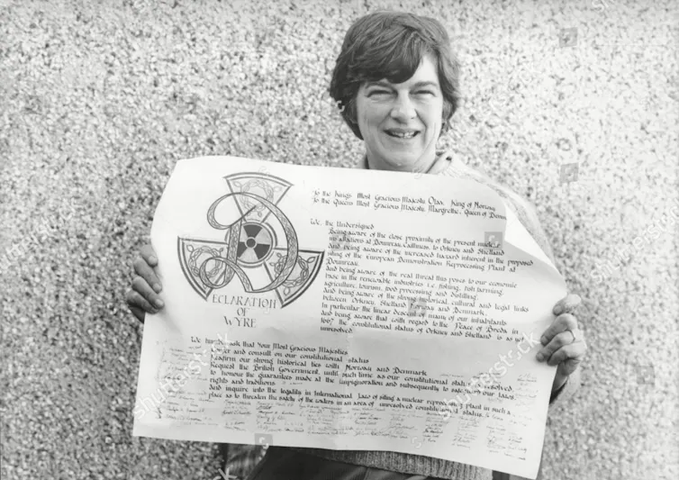A black and white image of a woman holding up a document standing against a wall.