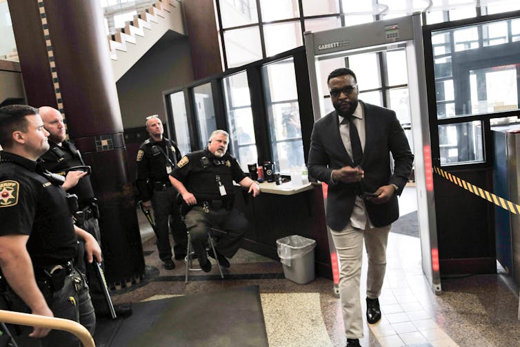 A black man wearing sunglasses walks through a metal detector as several white police officers watch him.