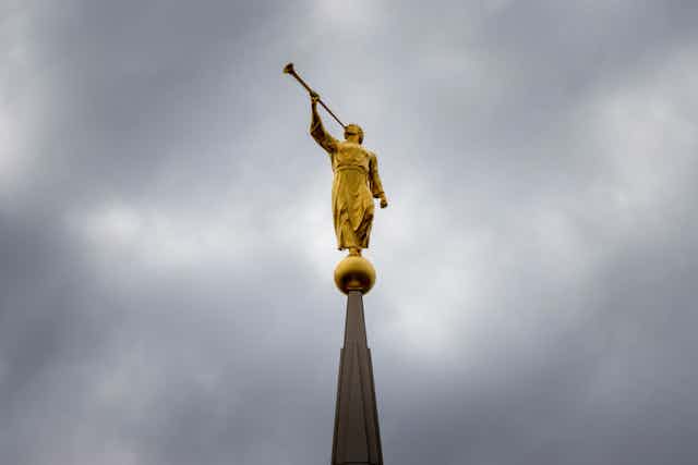 A golden statue of a man in a robe blowing a trumpet atop a steeple, seen against a gray sky.