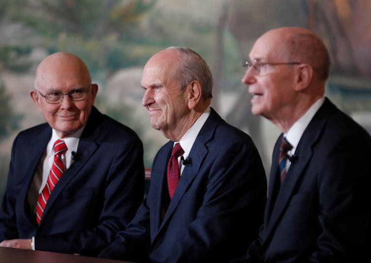 Three older men in suits and ties, with balding hair, sit while addressing an audience out of view.