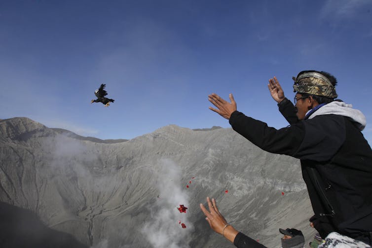 A group of people gather at a precipice, throwing flower petals and a chicken into the void.