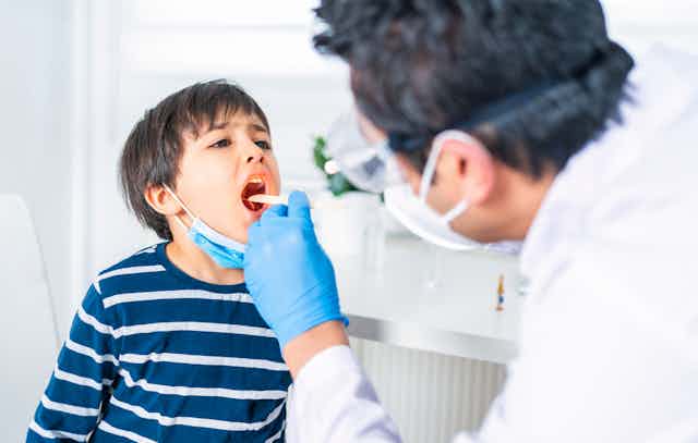 Male pediatrician leans forward to examine a young boy's throat with a tongue depressor at a clinic.