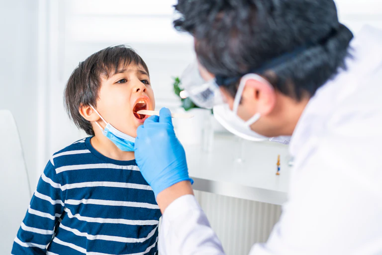 Male pediatrician leans forward to examine a young boy's throat with a tongue depressor at a clinic.