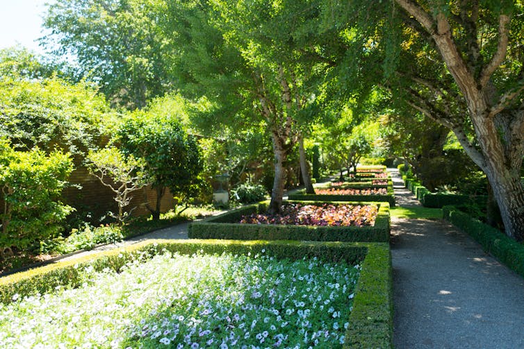 A garden walk with parallel gravel pathways on either side of carefully manicured flower beds and old trees.