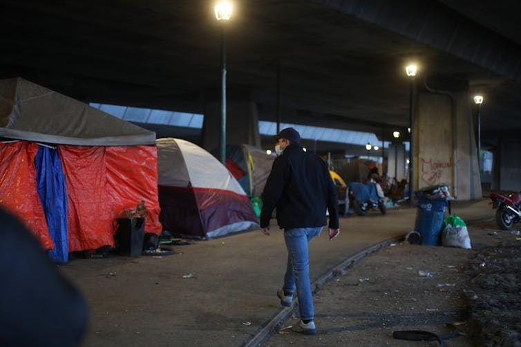 A man in a winter jacket, ball cap and face mask walks at night by tents in San Jose used by people who have no solid housing options.