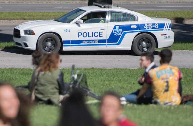 People sit in a park on a sunny day as a police cruiser is seen in the background.
