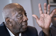 A Black bald man gestures as he speaks.