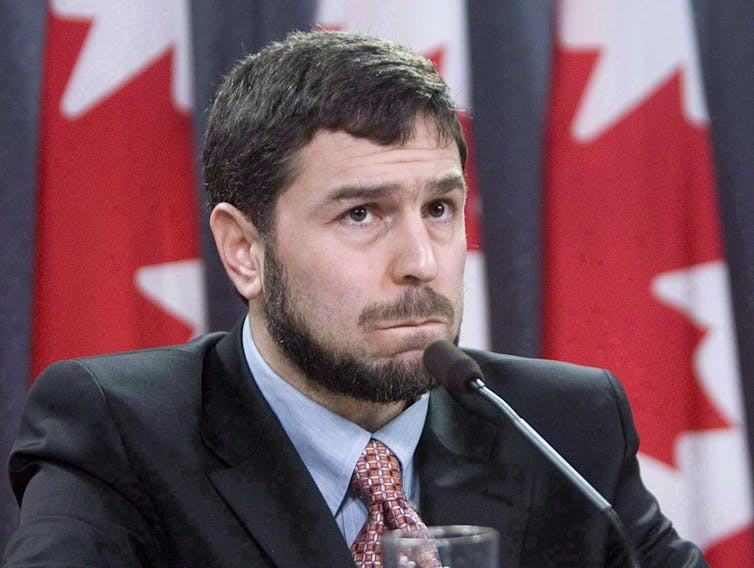 A bearded man listens to a question at a news conference. A row of Canadian flags is behind him.