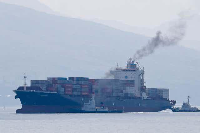 Exhaust smoke comes from container ship being escorted into port by tugs