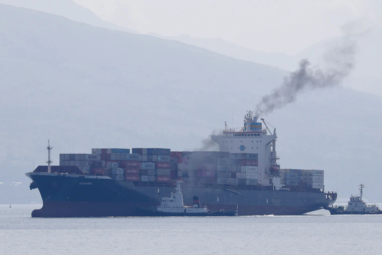 Exhaust smoke comes from container ship being escorted into port by tugs