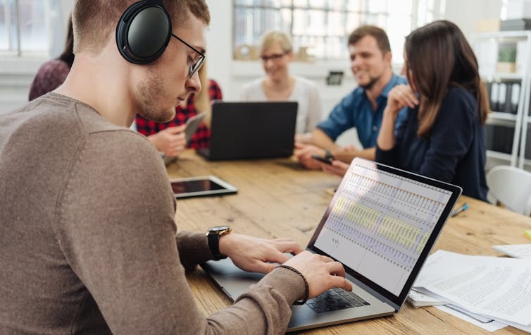 Man wearing headphone and typing on laptop while others around him talk.