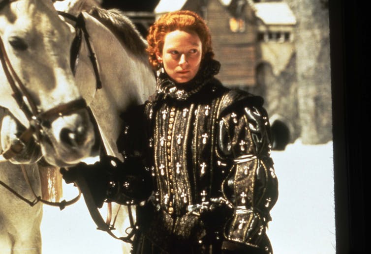 Una mujer andrógina vestida de época mira fuera de encuadre mientras sujeta las riendas de un caballo.