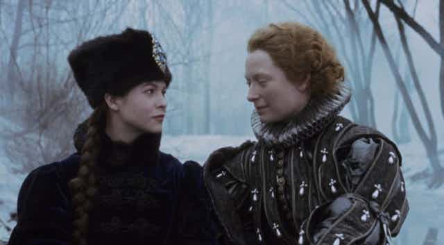 Una mujer con abrigo y gorro de pelo mira a otra frente a ella vestida de caballero del siglo XVII con cuello de lechuguilla.
