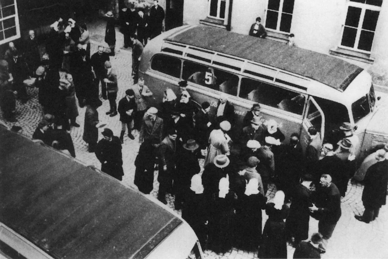 Una fotografía en blanco y negro de dos autobuses aparcados rodeados de figuras que van entrando en ellos.