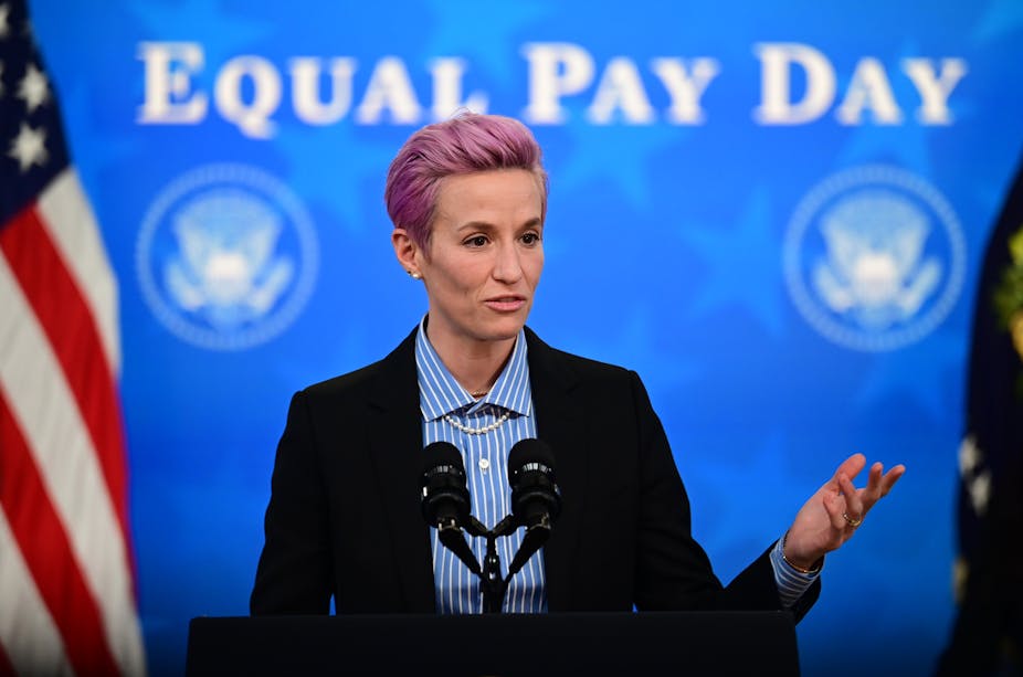 Megan Rapinoe prononce un discours devant un drapeau américain. Sur le mur derrière elle, l'inscription « Equal Pay Day »