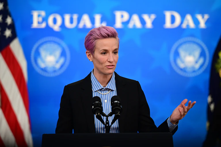 Megan Rapinoe prononce un discours devant un drapeau américain. Sur le mur derrière elle, l'inscription « Equal Pay Day »