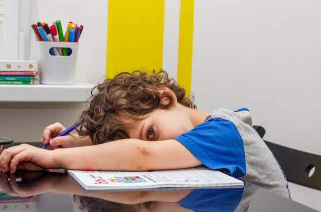 Un niño tumbado encima de una mesa y un cuaderno con deberes.