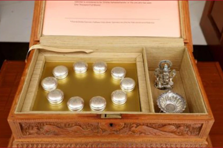 A wooden box with ten small silver containers with an image of a Ganesha and a bowl in a side compartment.