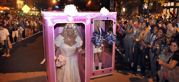 Two drag queens march down a street wearing pink Barbie boxes to create the effect of being dolls before crowds of onlookers.