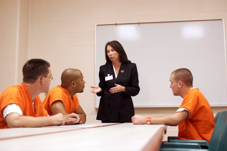 Standing before a whiteboard, a female instructor talks to three prisoners seated at a table and dressed in orange jump suits.