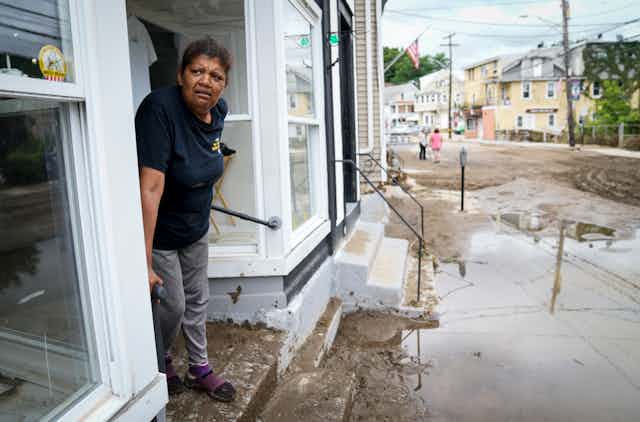 A woman stands on a storefront stoop that is covered in mud. The street is filled with mud and water. American flags hang on the poles outside.