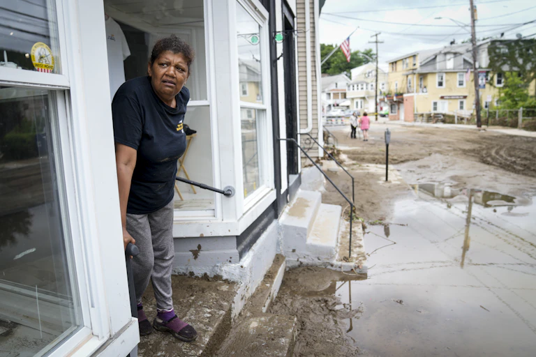 A woman stands on a storefront stoop that is covered in mud. The street is filled with mud and water. American flags hang on the poles outside.