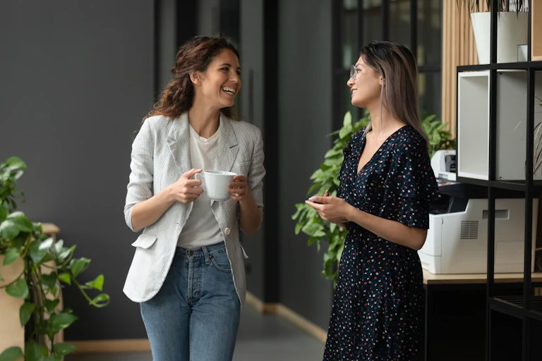 Two women in workplace attire have a conversation while standing in an office