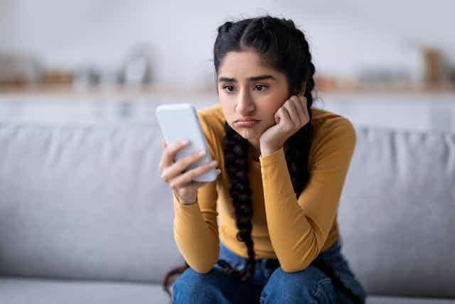 A photo of a young woman sitting on a couch staring at her phone with a sad expression on her face.