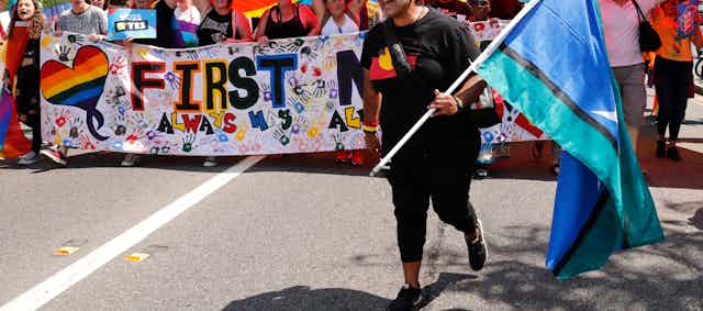 A person walks in front of a large banner carrying a Torres Strait Islander flag, and wearing an Aboriginal flag t shirt.