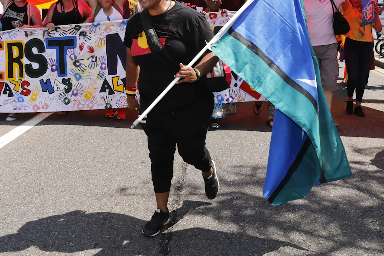 A person walks in front of a large banner carrying a Torres Strait Islander flag, and wearing an Aboriginal flag t shirt.