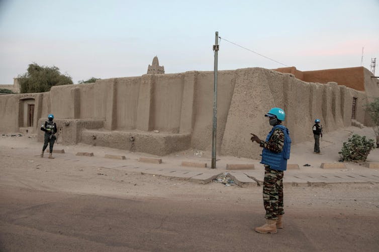 Three men in military uniforms and blue helmet standing outside the walls of a building on a quiet street.