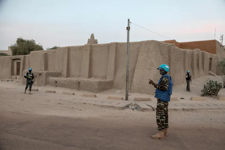 Three men in military uniforms and blue helmet standing outside the walls of a building on a quiet street.
