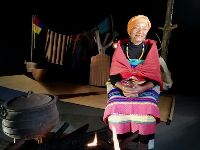 An elderly woman sits in traditional clothing on a stage setting of a rural homestead. Her hands in her lap, she smiles at the camera.