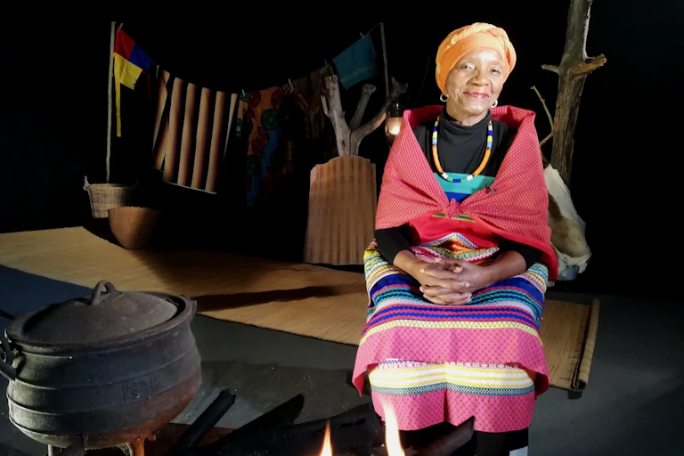 An elderly woman sits in traditional clothing on a stage setting of a rural homestead. Her hands in her lap, she smiles at the camera.