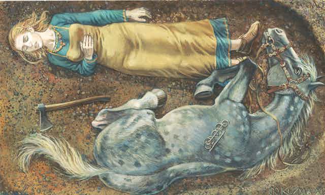 A blonde woman lies on her back in a blue and gold dress. She shares her grave with a grey horse and an axe lays between then. 
