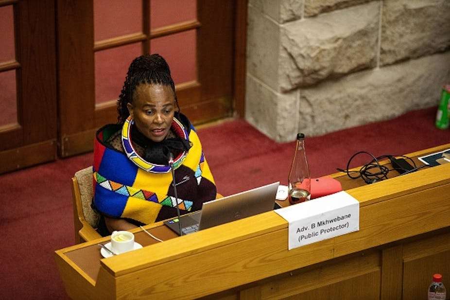A woman with plaited hair, dressed in traditional Ndebele garb, sits behind a desk with a laptop open, answering questions at a hearing.