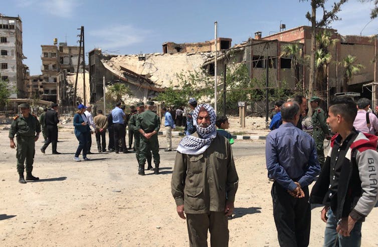 Police officers and others stand in front of tall buildings that appear to be damaged.