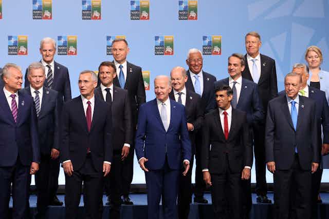 A group of mostly men in business attire, including Joe Biden and Emmanuel Macron, stand next to each other. 