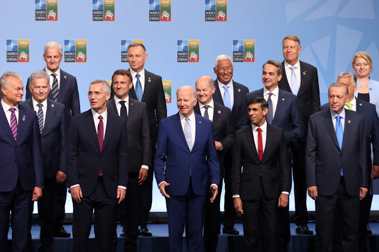 A group of mostly men in business attire, including Joe Biden and Emmanuel Macron, stand next to each other.