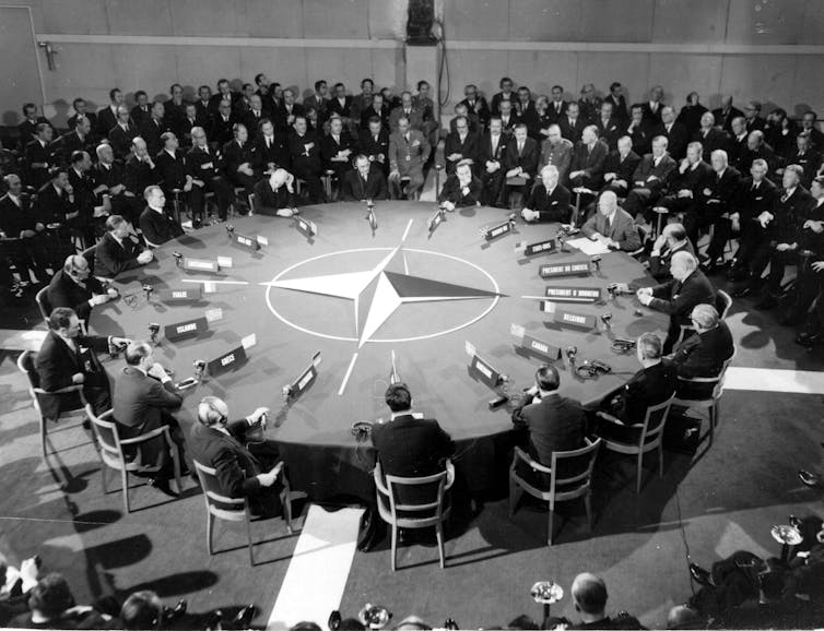 People sit around a large circular table with a compass on it in a black and white photo.