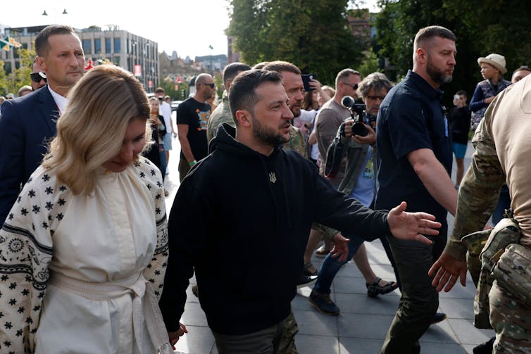 Volodymr Zelenskyy walks through a crowd with his wife, surrounded by men in suits.
