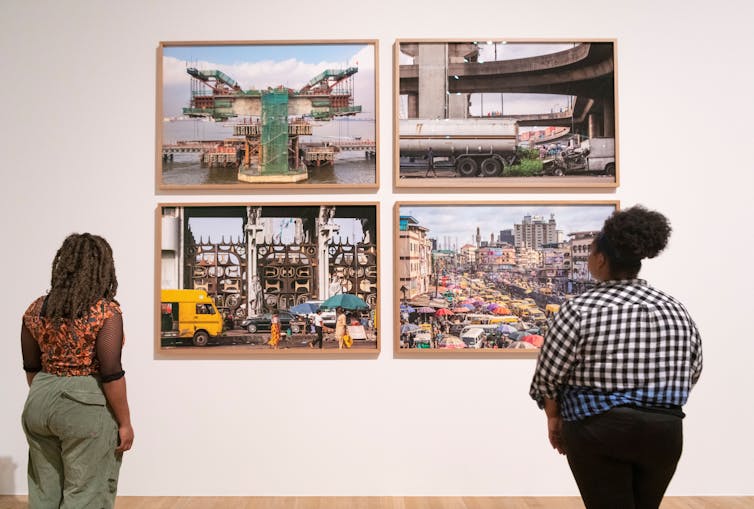 Two black women looking at four photographs on a blank wall showing urban life.