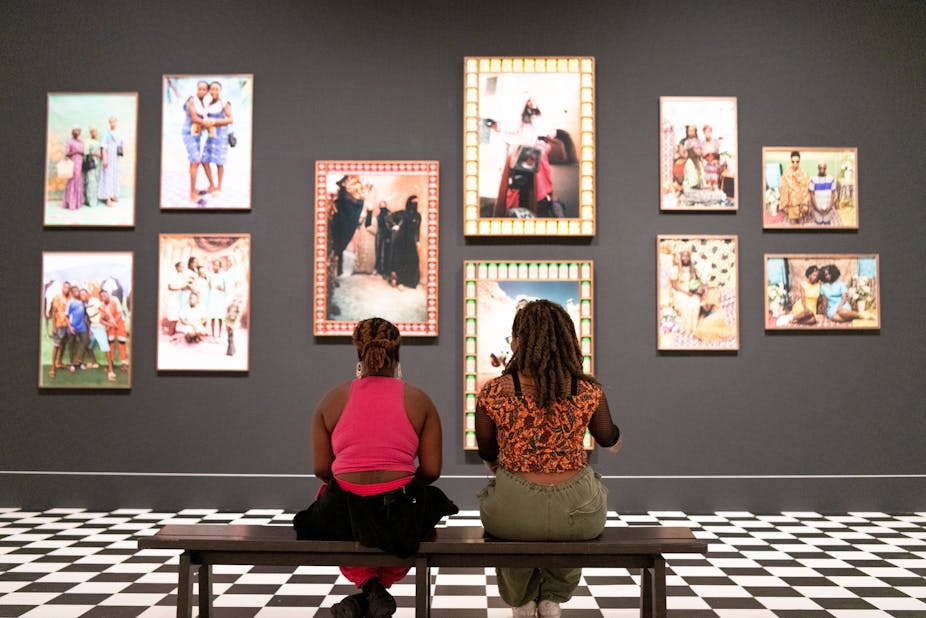 Two black women sat on a bench looking at large, colourfully framed photographs hung on a grey wall. 