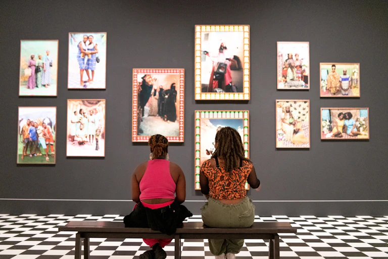 Two black women sat on a bench looking at large, colourfully framed photographs hung on a grey wall.
