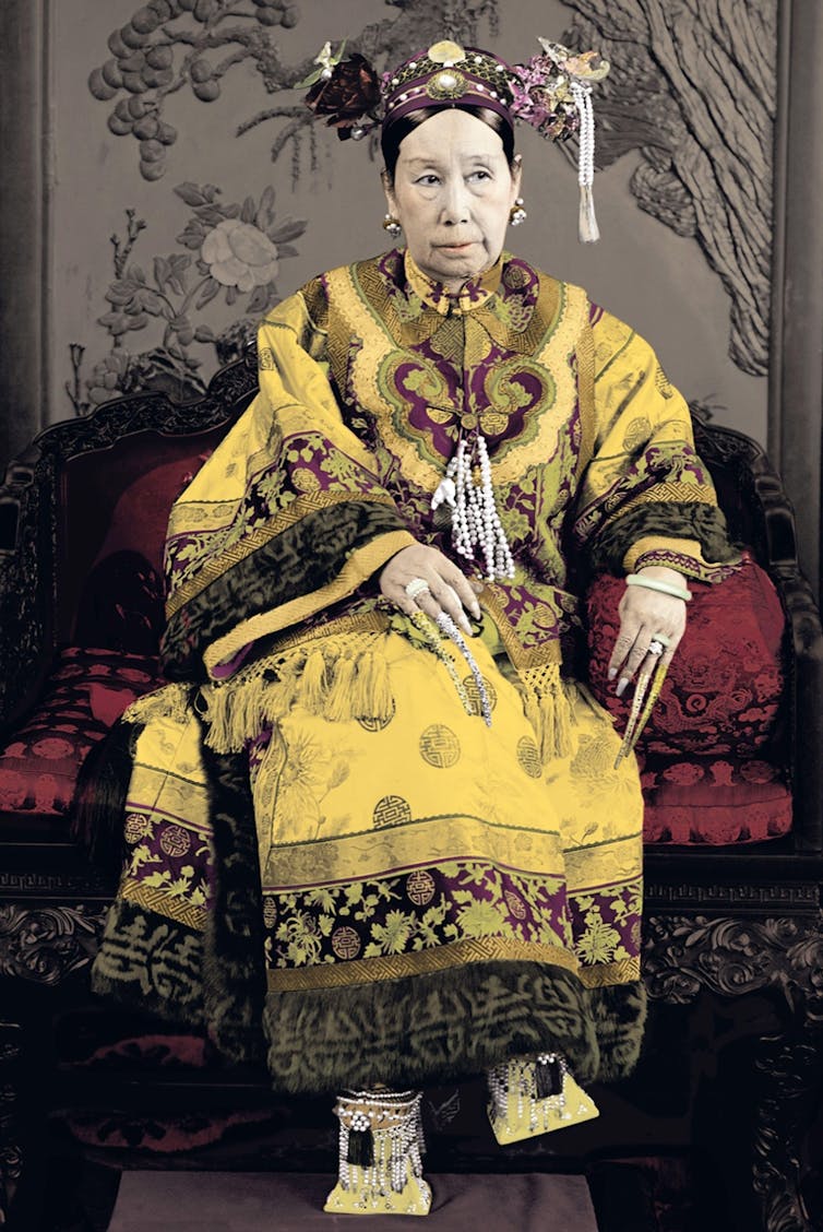 A Chinese woman dressed in yellow robes sitting on a red banquette.